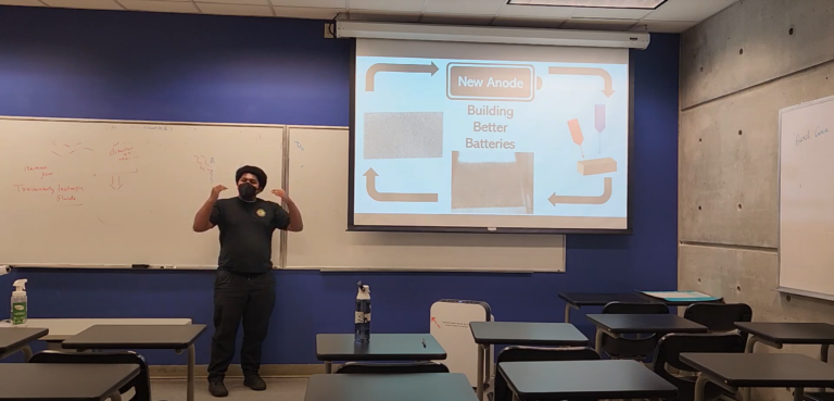 Adult African-American man wearing a black mask talks about presentation titled "Building Better Batteries".